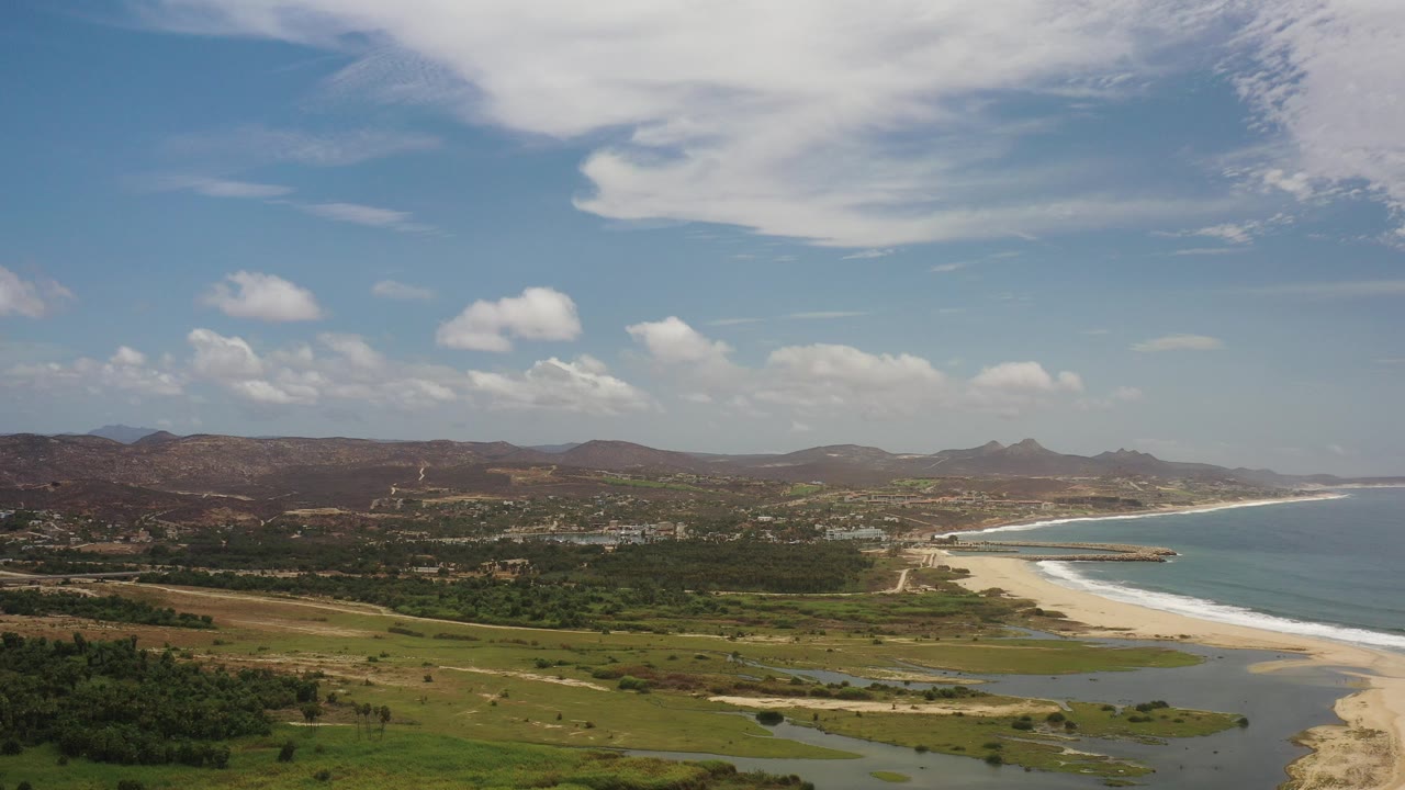 Opening frame of Los Cabos, Mexico coastline with a curved sandy beach, blue sea to the right, green lowland and water channels in the foreground, brown hills in back, and no visible people.