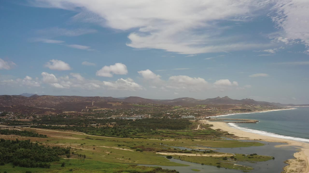 Los Cabos, Mexico aerial view with the beach arc slightly farther along the frame, blue water meeting pale sand, green foreground vegetation, distant hills, and no visible people.