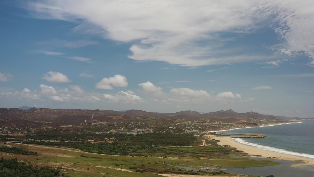 Opening wide view of the Los Cabos, Mexico coastline, with green lowlands in front, a pale beach curving toward blue water, distant hills, scattered buildings, and no visible people.