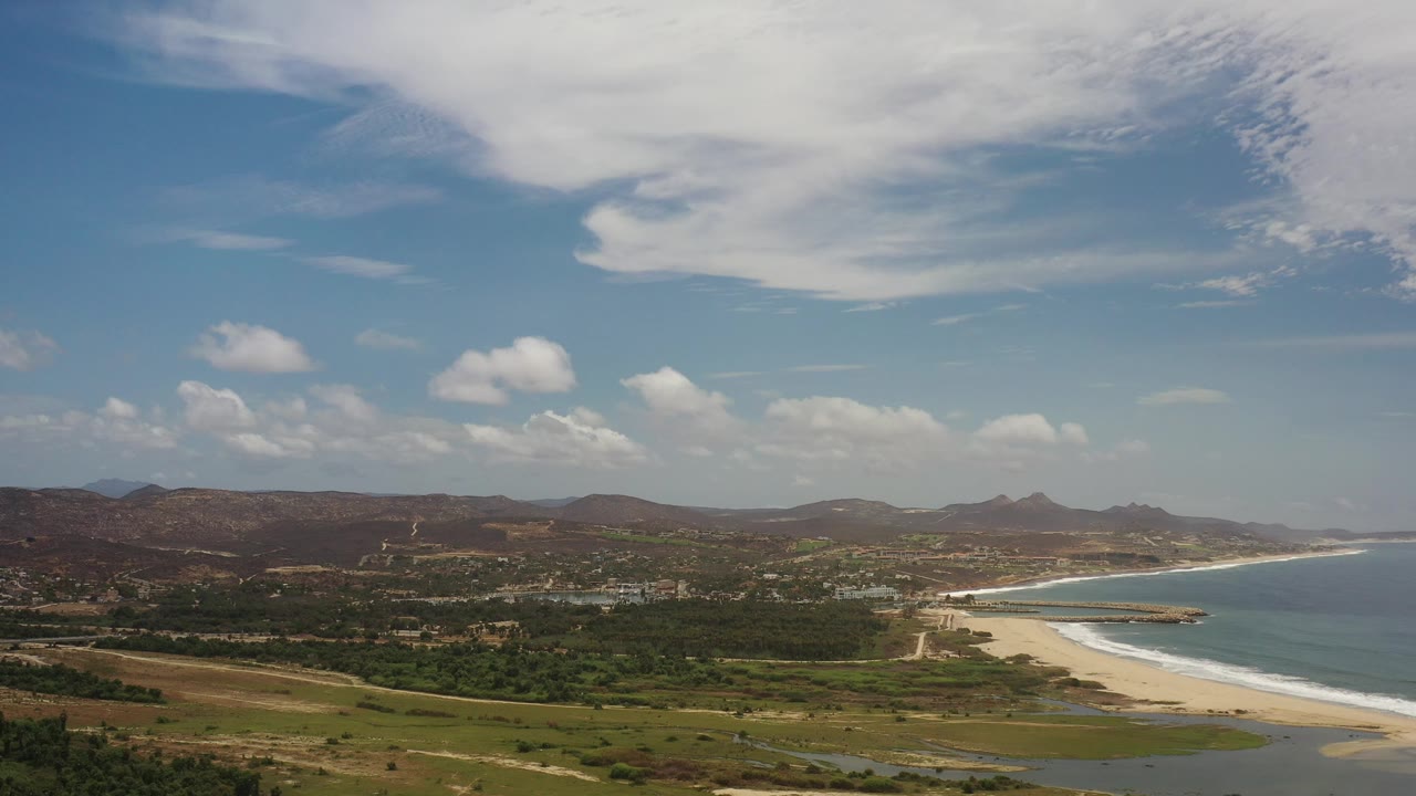 Coastal panorama with long beach, ocean, greenery, distant hills, and bright sky.