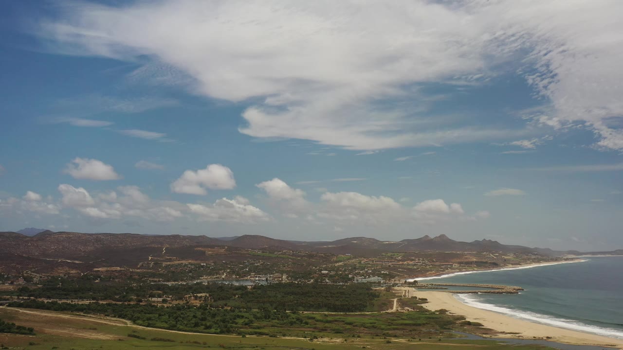 Second frame shows the Los Cabos beach curve and calm sea to the right, with green lowland, dry hills, scattered buildings, large overhead cloud cover, and no visible people.