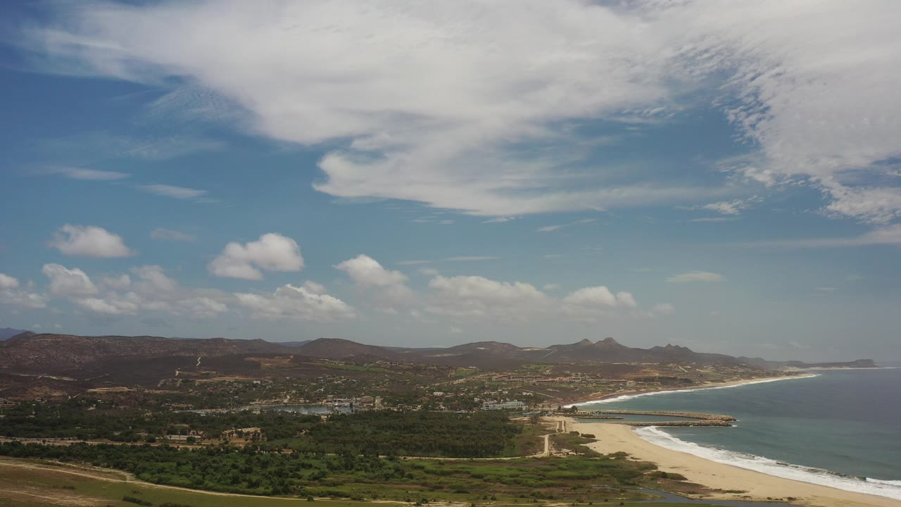Wide aerial coast with green land, sandy beach, ocean, hills, and clouded sky.