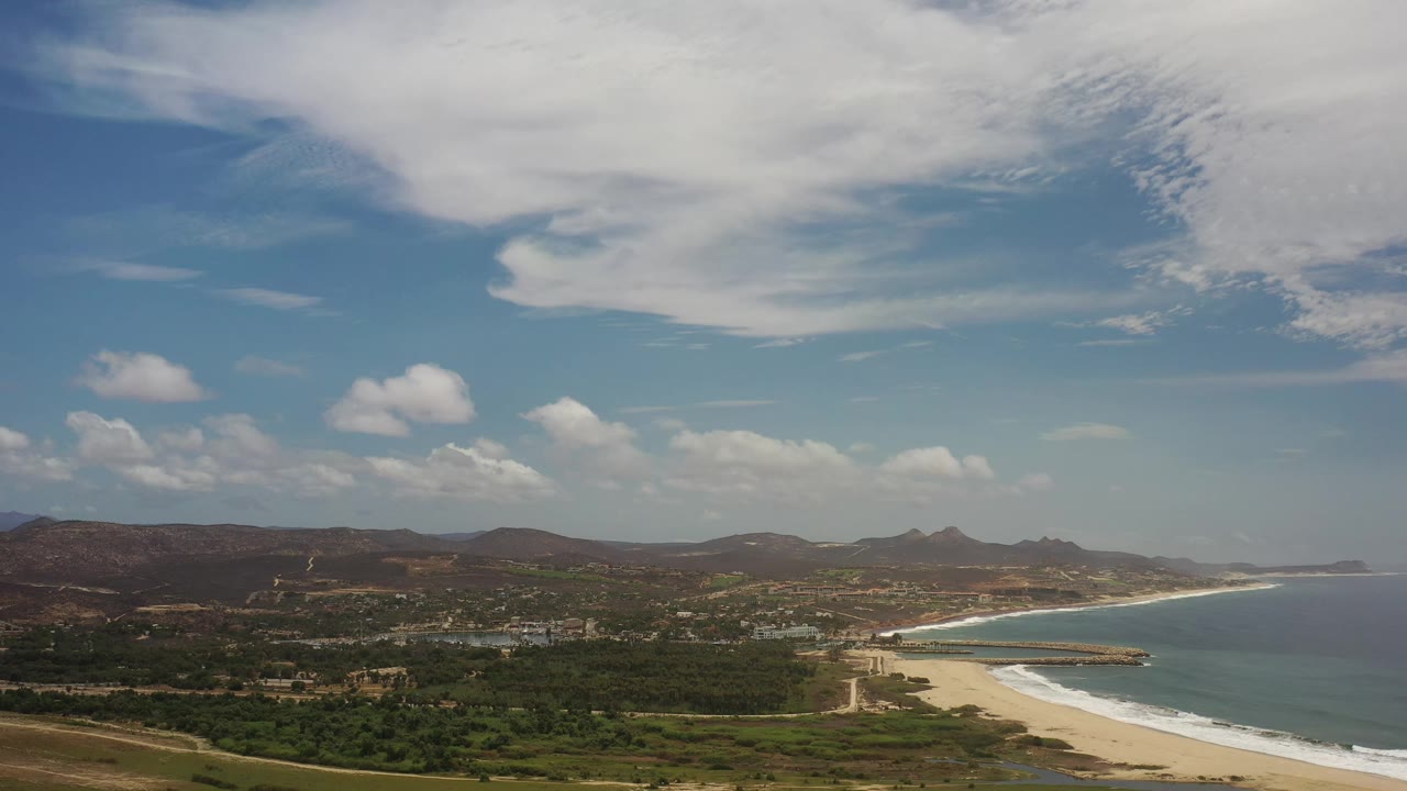 Aerial frame over Los Cabos, Mexico showing green foreground terrain, a long beach and surf band on the right, ocean water, brown hills, bright sky, and no visible people.