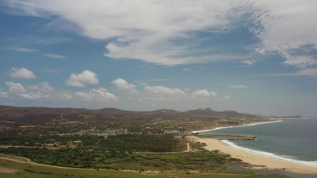 Wide aerial view over the Los Cabos coastline with a pale sand beach, blue-green ocean, inland hills, scattered buildings, and no visible people.