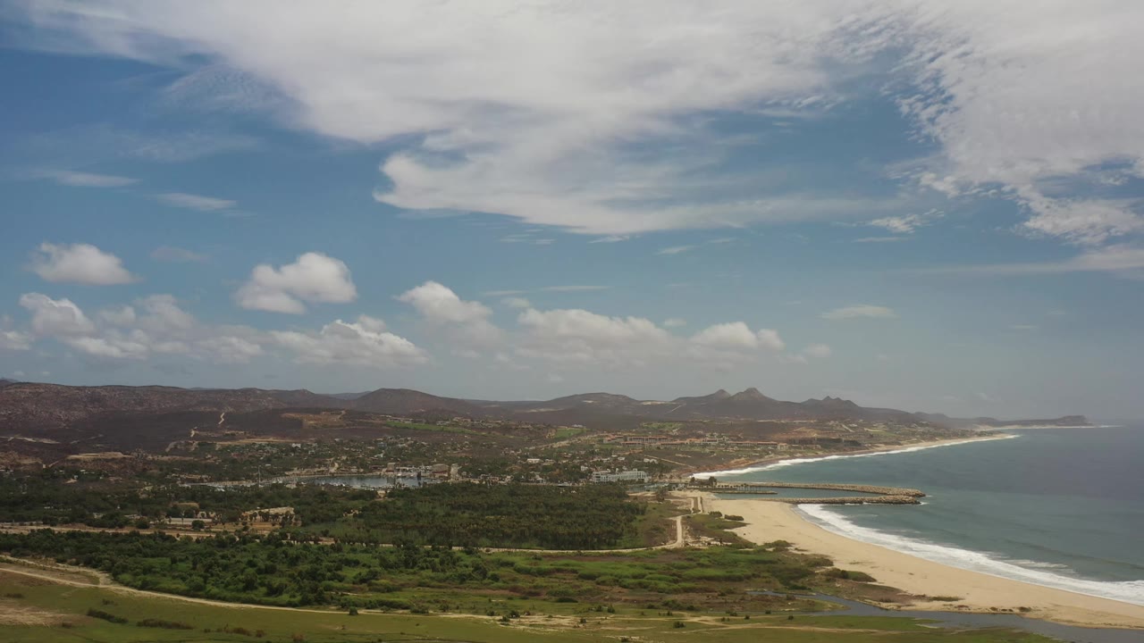 Los Cabos aerial frame showing the beach curve tighter to the right, white surf along the sand, green lowland vegetation, distant hills, and no visible people.