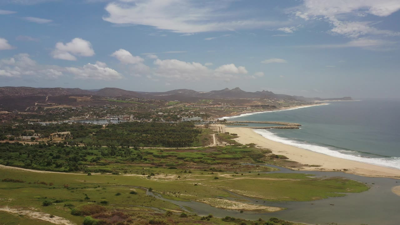 Opening aerial frame over Los Cabos, Mexico with inland tidal pools in the foreground, a pale beach and surf line at midframe, scattered development beyond the sand, distant dry hills, and no visible people.