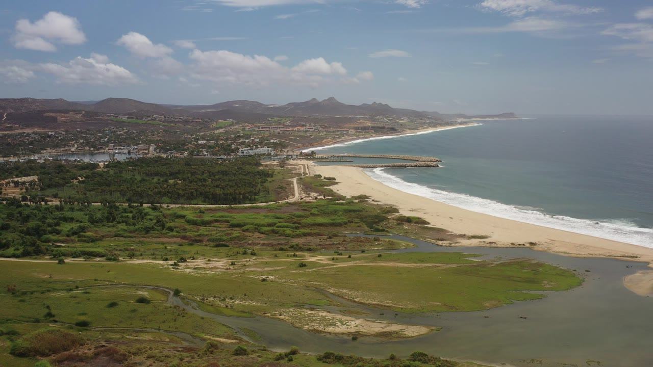 Wide aerial view over Los Cabos, Mexico, with green lagoon flats in the foreground, a pale sandy beach, white surf, blue ocean, distant hills, and no visible people.