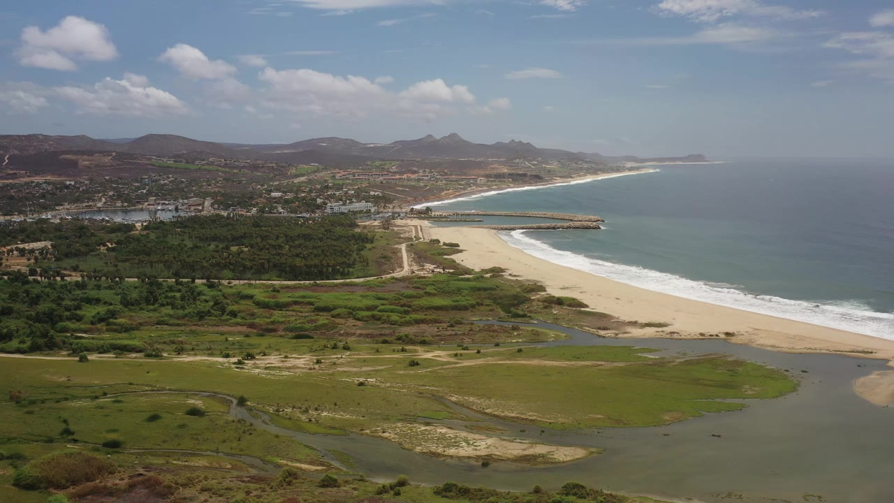 Aerial frame over Los Cabos, Mexico, showing a curved beach and surf line beside green lowlands, with town structures and brown hills under scattered clouds, and no visible people.