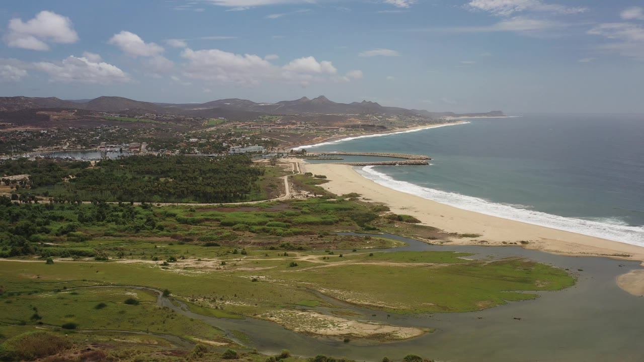 Drone view of sandy coastline, green flats, ocean, and mountains.