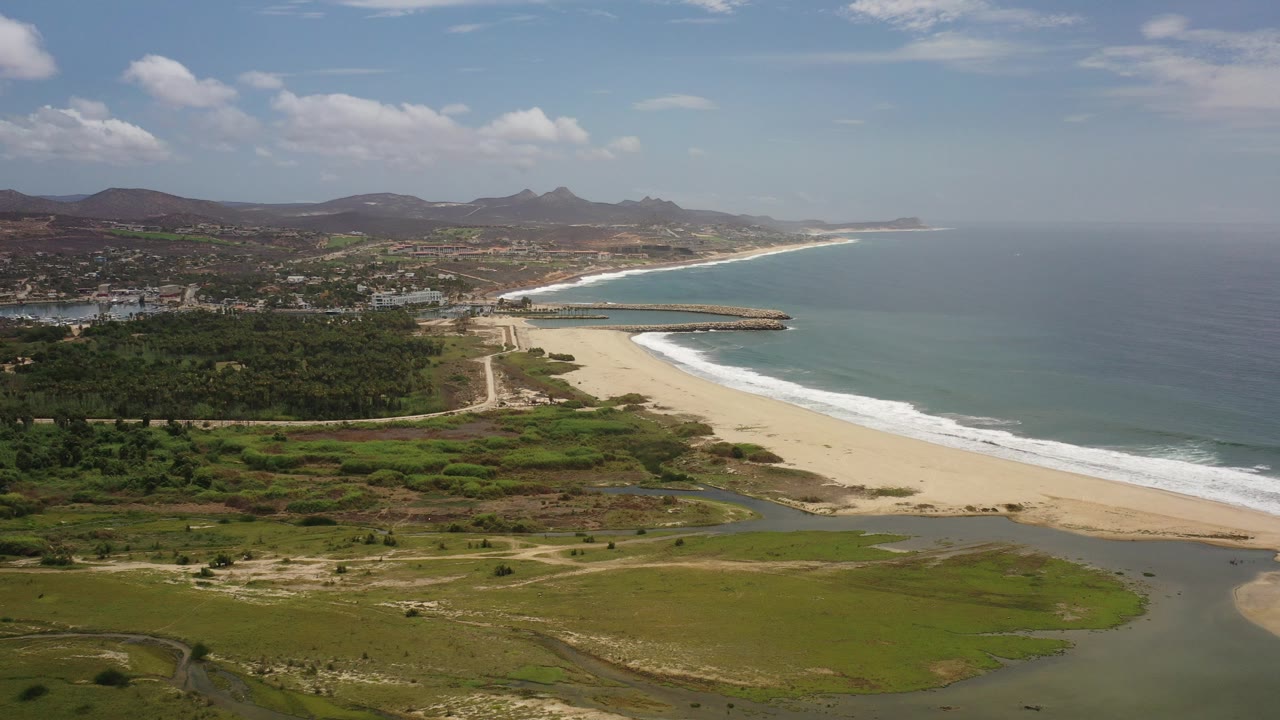 Wide aerial view in Los Cabos, Mexico showing a sandy beach beside blue-green ocean, a lagoon in the foreground, green scrubland, distant buildings, hills, and no visible people.
