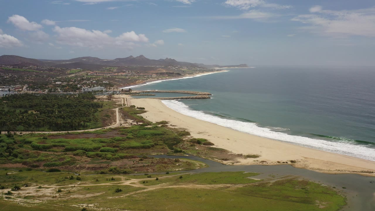 Wide aerial frame over the Los Cabos, Mexico coast showing pale sand, white surf, blue-green ocean, scrubby foreground wetlands, distant hills, and no visible people.
