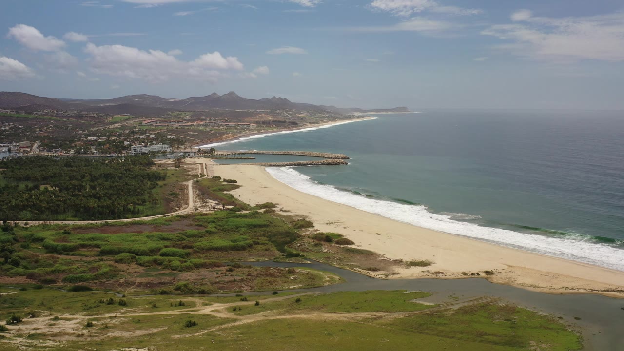 Drone-like view of shoreline, waves, breakwater, town edge, and hills.