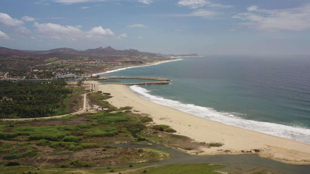 Aerial view in Los Cabos, Mexico with pale beach curving along blue-green ocean, white surf, inland greenery, distant hills, and no visible people.