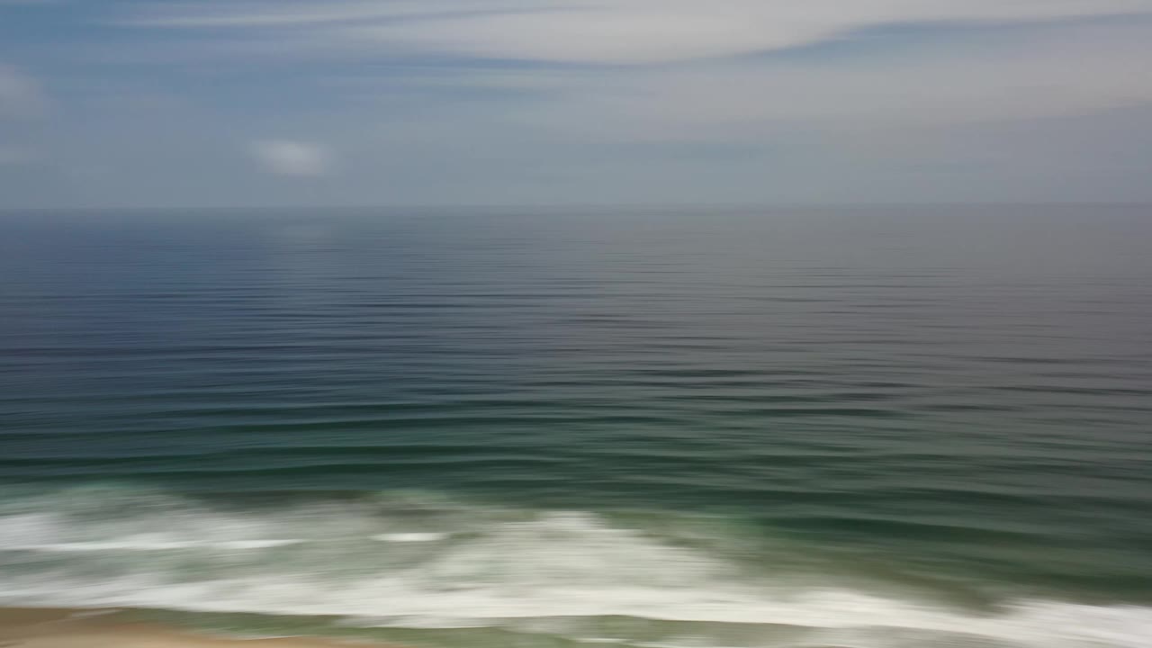 Aerial coastal frame in Los Cabos, Mexico showing a level ocean horizon, repeated swell bands, white breaking surf along the lower frame, and no visible people.