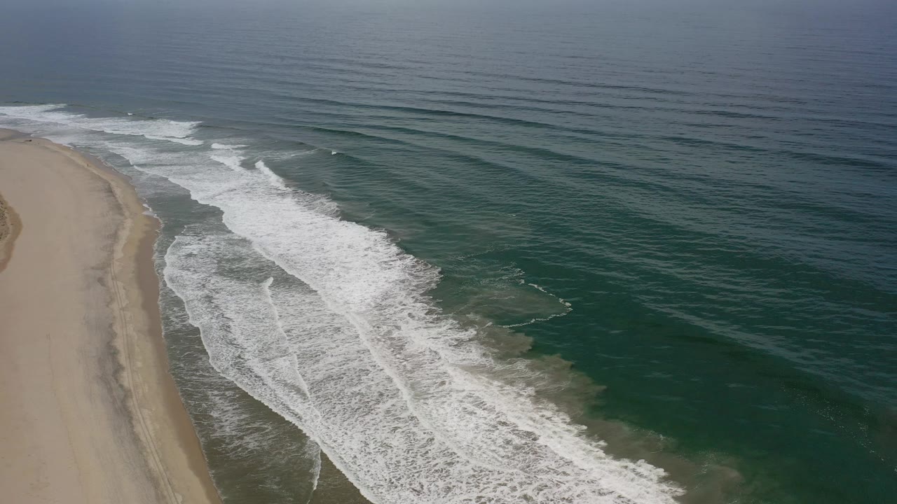 Aerial frame over the Los Cabos coastline with a sandy beach on the left, green water on the right, white surf crossing diagonally, and no visible people.