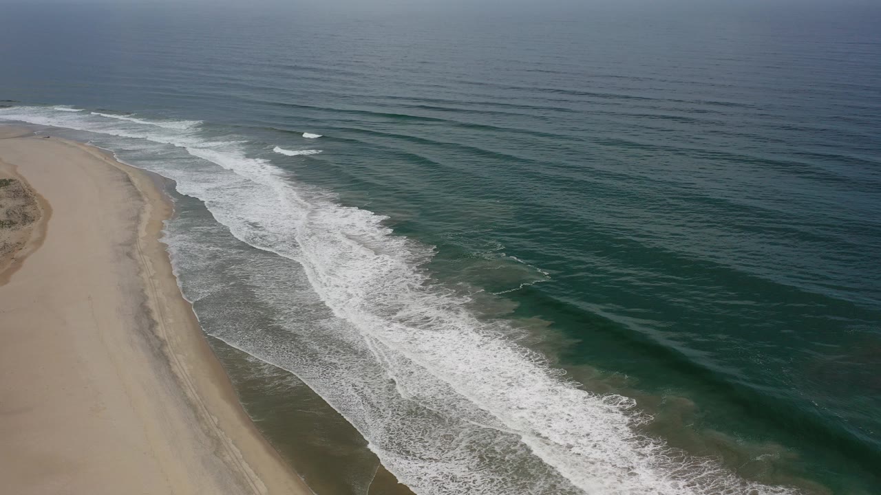 Early frame of a Los Cabos, Mexico coastline with sandy beach on the left, teal ocean on the right, white surf crossing diagonally, and no visible people.