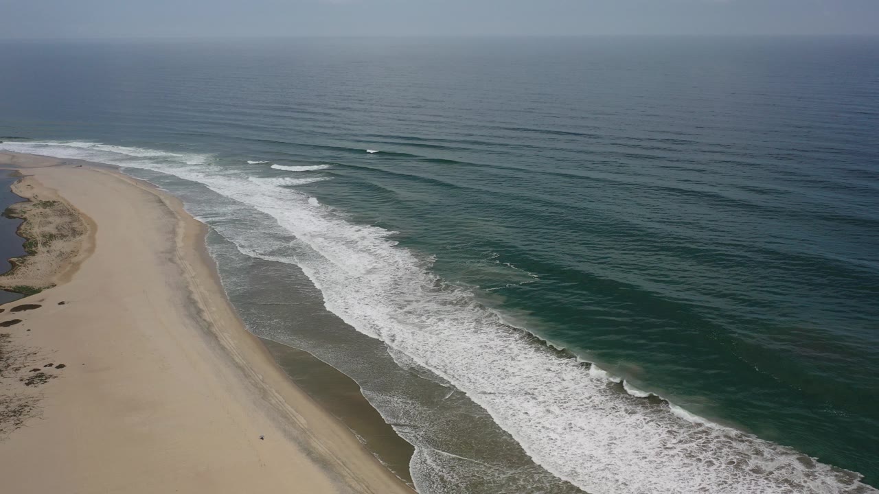 Elevated aerial view in Los Cabos, Mexico, showing a pale sandy beach curving along dark blue-green surf with a hazy horizon and no visible people.