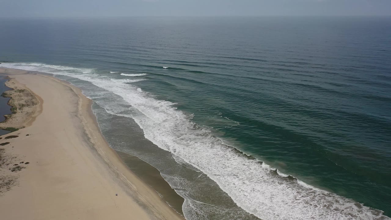 Aerial frame of the Los Cabos shoreline with long white foam lines crossing the midground, open ocean beyond, and no visible people on the sand.