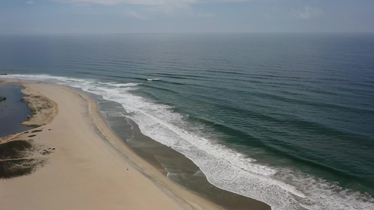 Opening aerial frame over a Los Cabos beach, with tan sand on the left, white surf bands near shore, dark blue-green ocean to the right, and no visible people.