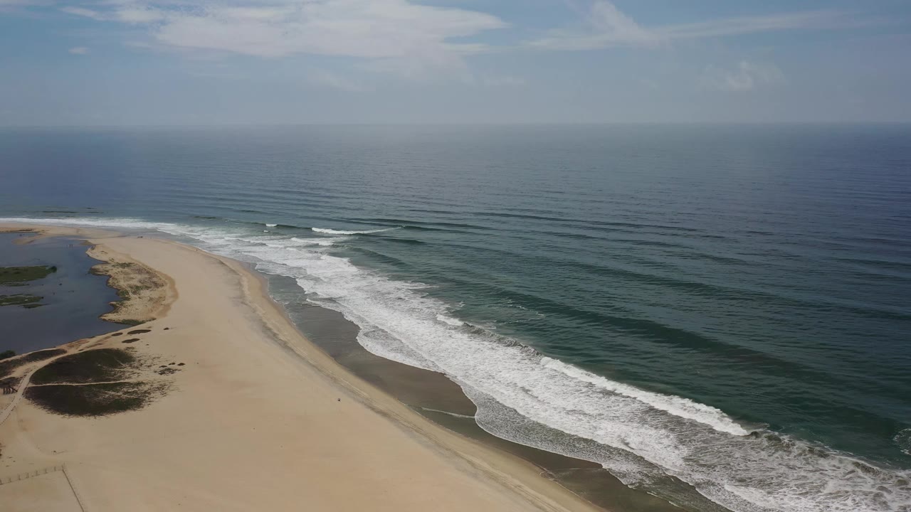 Aerial Los Cabos coastline frame with the beach curve more centered, white foam bands along the right, calm dark water offshore, dune scrub below, and no visible people.