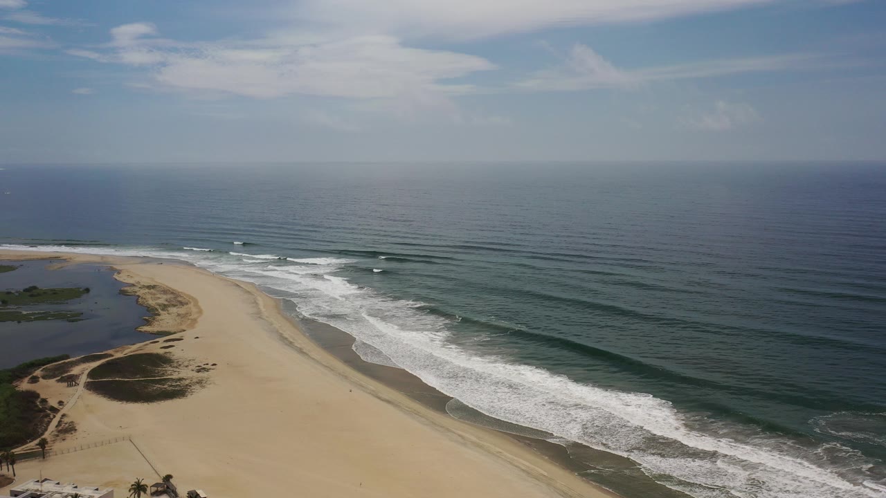 Aerial view over a Los Cabos beach with a lagoon on the left, white surf meeting wide sand, dark ocean beyond, and no visible people.