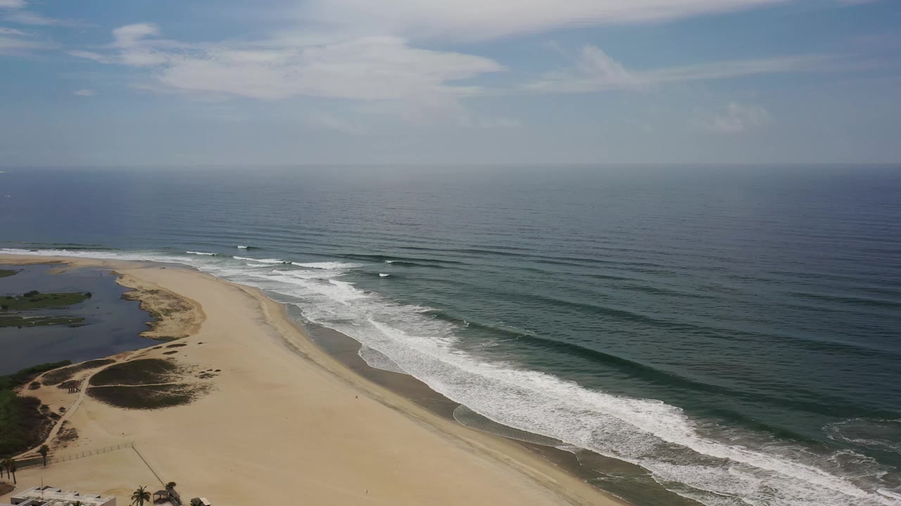 High aerial frame of the Los Cabos shoreline, with parallel wave bands rolling toward pale sand and a lagoon edge visible to the left, no visible people.
