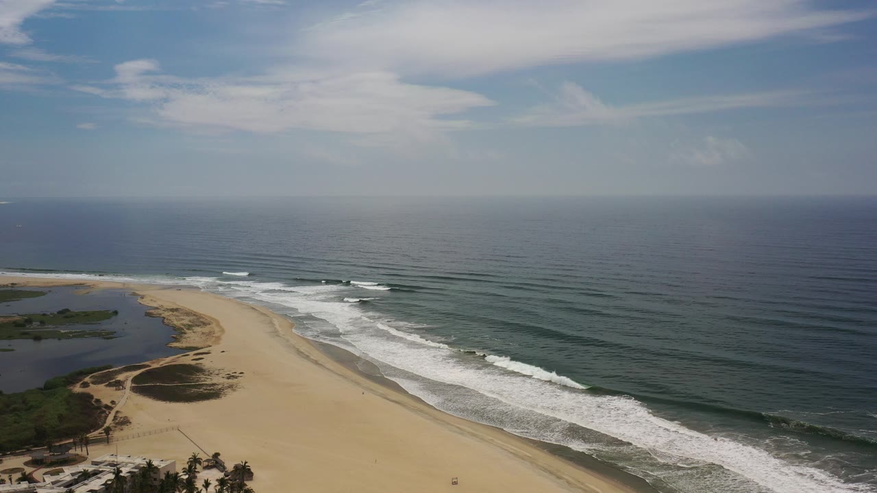 High aerial view over Los Cabos, Mexico, with a wide sandy beach curving between a lagoon on the left and breaking surf on the right, with no visible people.