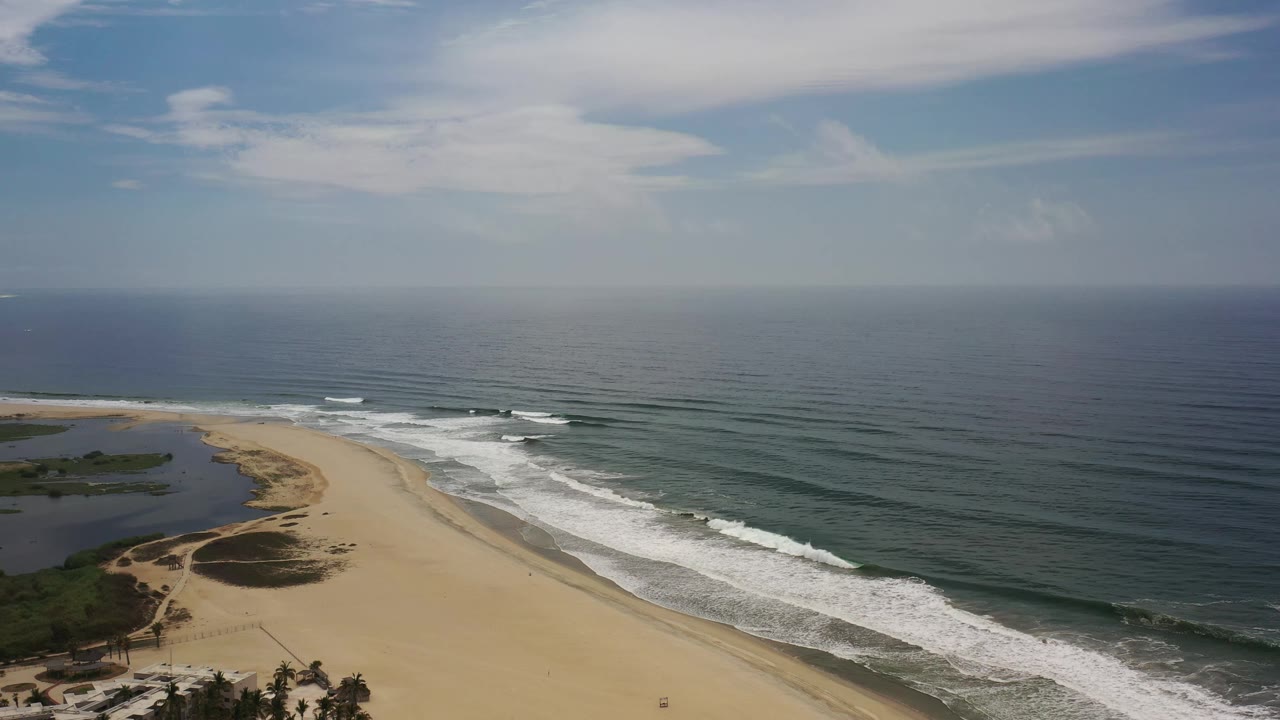 Aerial Los Cabos beach frame with the lagoon more centered at left, white surf bands rolling along the shoreline, blue-green ocean beyond, and no visible people.