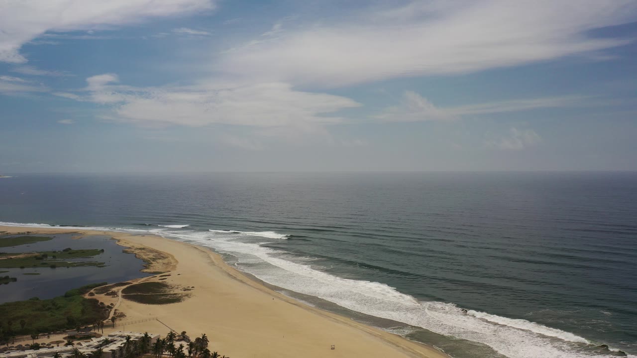 Wide aerial view of a Los Cabos beach with a sandy curve, surf bands, ocean horizon, lagoon at left, and no visible people.