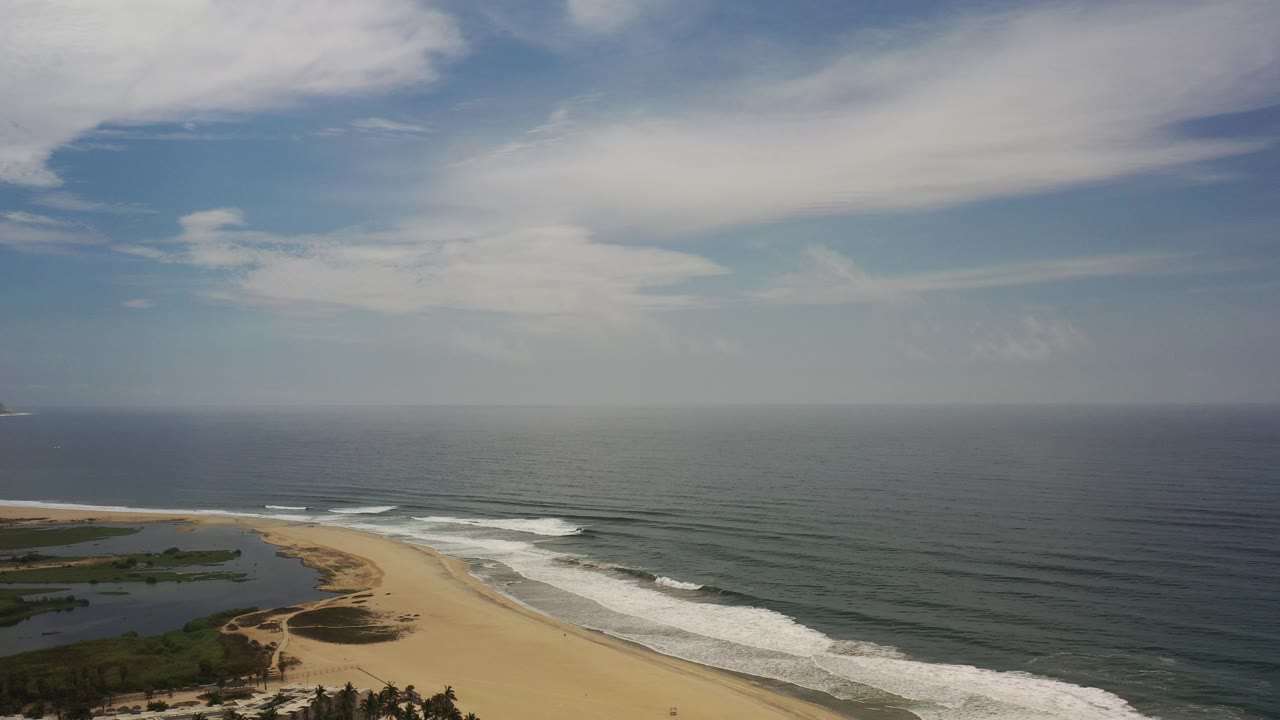 Opening aerial view over a Los Cabos beach, with pale sand at lower left, white surf bands, dark ocean to the horizon, high clouds, and no visible people.