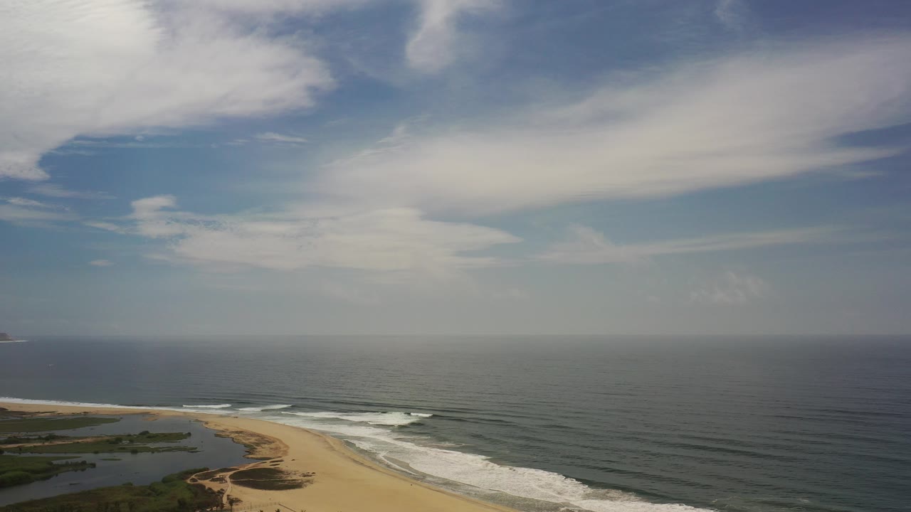 Aerial coastline with surf, sand, ocean, and cloudy sky