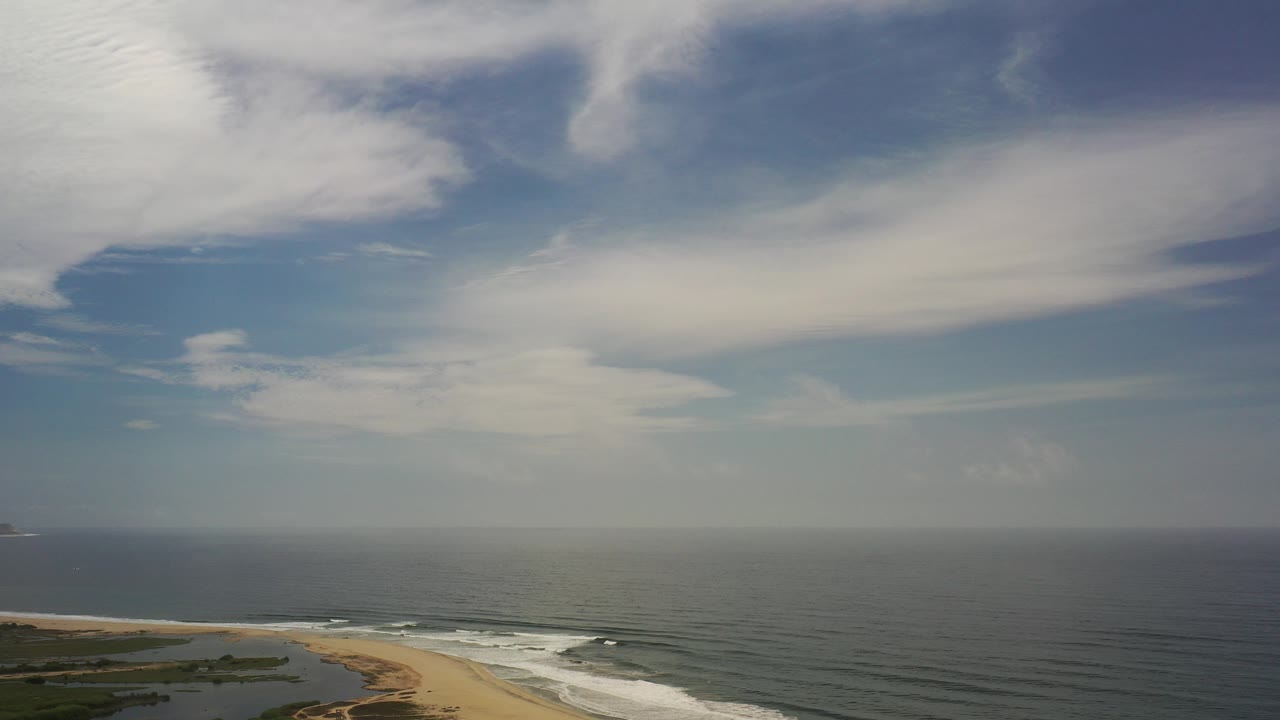 Aerial coastline with sandy beach, surf, ocean, and broad clouds.