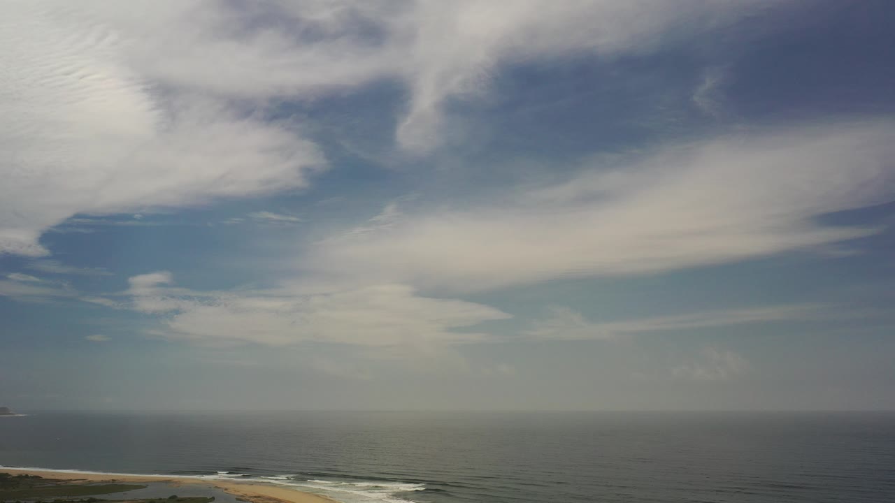 Wide aerial view in Los Cabos, Mexico, showing blue-gray sea below a cloud-filled light-blue sky, a thin beach edge at the bottom, and no visible people.