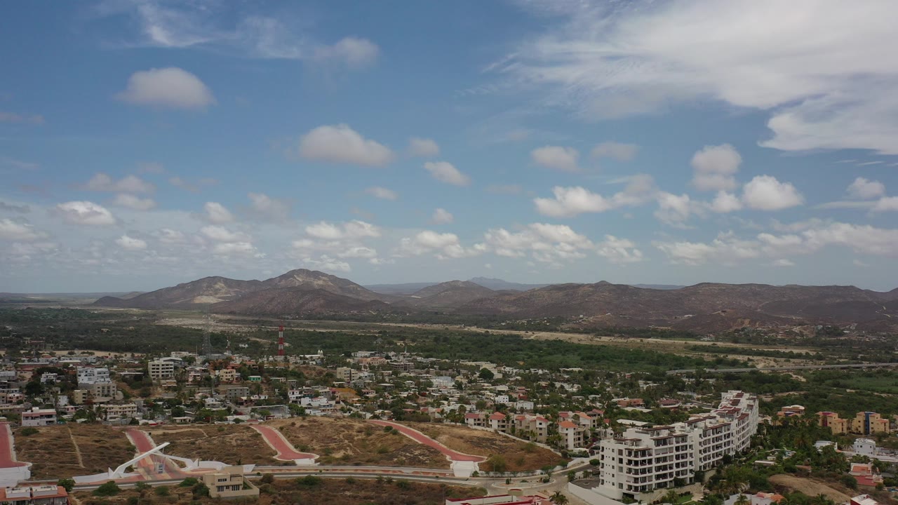 Opening frame of Los Cabos, Mexico from above, with town buildings at the bottom, dry hills and mountains across the horizon, scattered clouds in blue sky, and no visible people.
