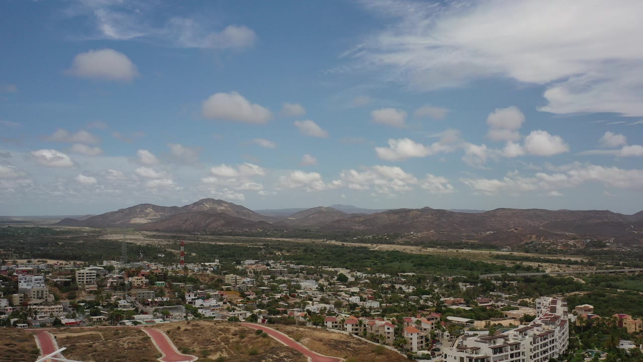 Wide aerial view over Los Cabos with low-rise buildings, dry hills, distant mountains, scattered clouds, and no visible people.