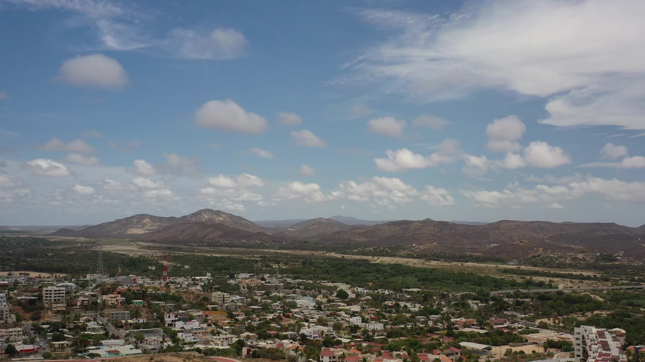 Opening aerial frame over Los Cabos, Mexico with broad blue sky, scattered white clouds, distant low mountains, green trees, and no visible people.