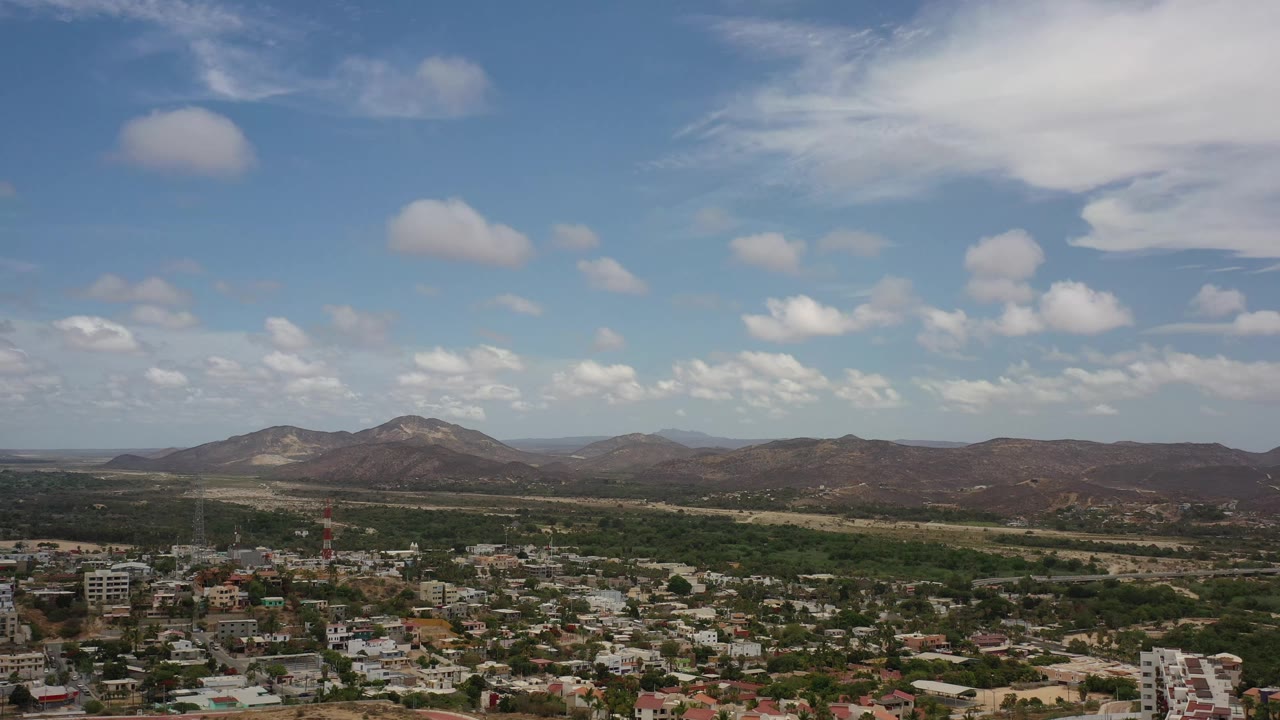 Early frame of Los Cabos, Mexico showing soft white clouds over brown mountain ridges, a green belt of trees, and clustered buildings with no visible people.