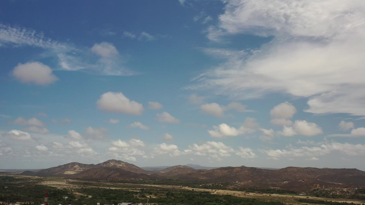 Opening aerial frame over Los Cabos, Mexico, with brown mountain ridges low in frame, a broad blue sky above, and no visible people.
