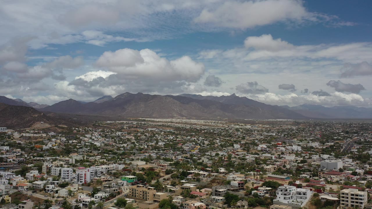 Los Cabos aerial view one second in, with the urban area spread across the lower frame, dark hills to the left, distant mountain ridges, and no visible people.