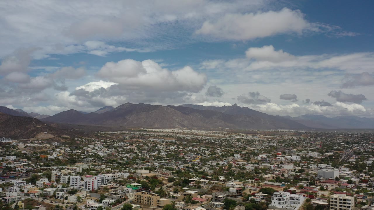 Opening aerial frame over Los Cabos, Mexico, with low-rise city blocks below, dark mountains on the horizon, broad white clouds overhead, and no visible people.