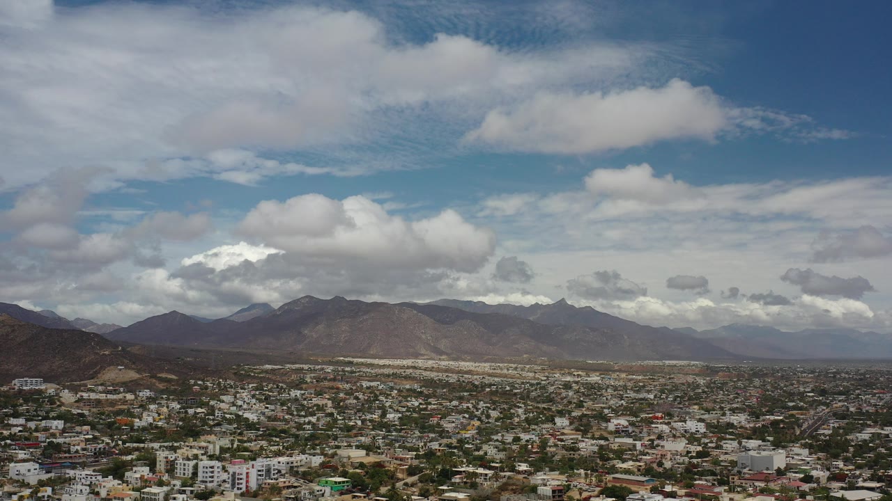 Opening frame of Los Cabos, Mexico, from an elevated view over low-rise buildings toward brown mountain ridges and a clouded blue sky, with no visible people.