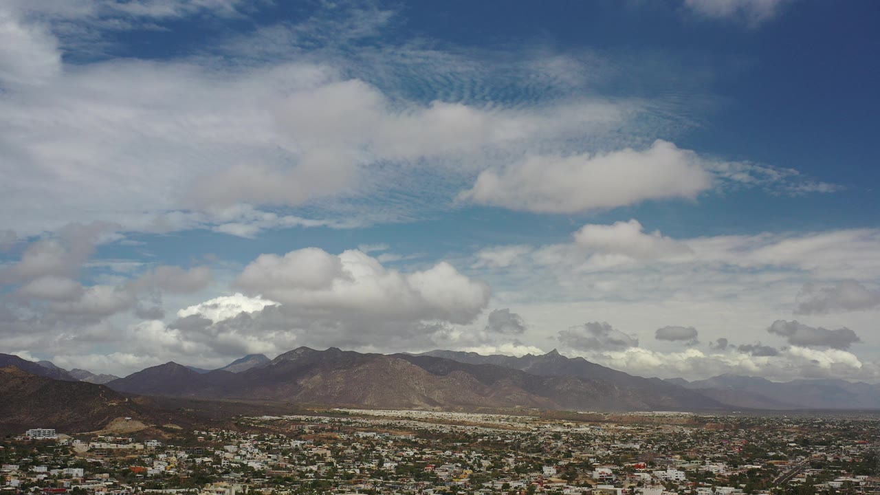 Opening frame of Los Cabos, Mexico from an elevated viewpoint, with distant rooftops at the bottom, brown mountain ridges on the horizon, layered clouds overhead, and no visible people.