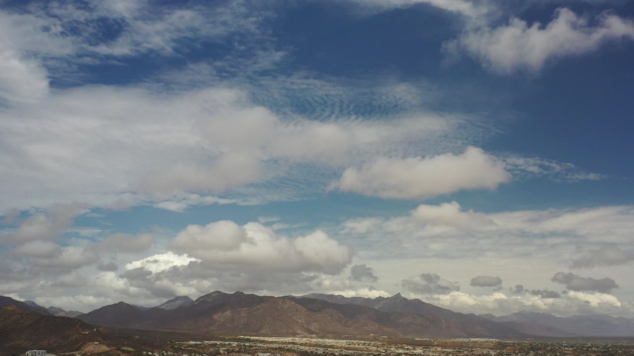 At 0:00, white and gray clouds spread across a blue sky above a distant Los Cabos mountain range, with no visible people.