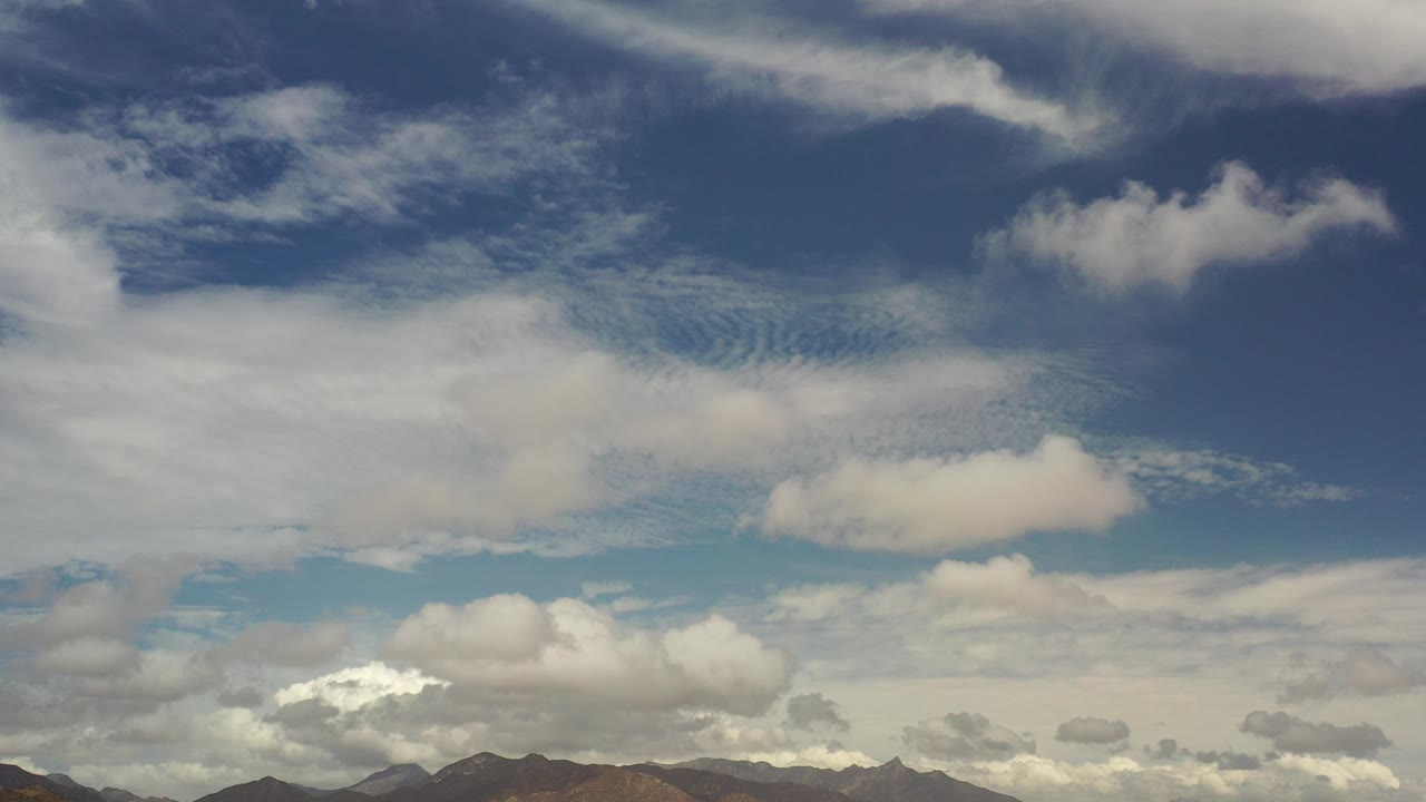 Opening frame of a blue Los Cabos, Mexico sky with broad white cloud layers, a thin mountain horizon at the bottom, and no visible people.