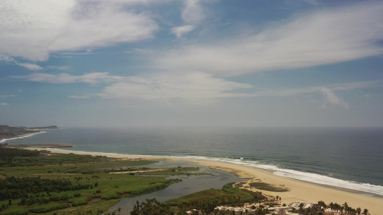 Wide aerial view over Los Cabos, Mexico, showing green coastal land, a lagoon mouth, pale sand, blue ocean surf, distant shoreline, and no visible people.