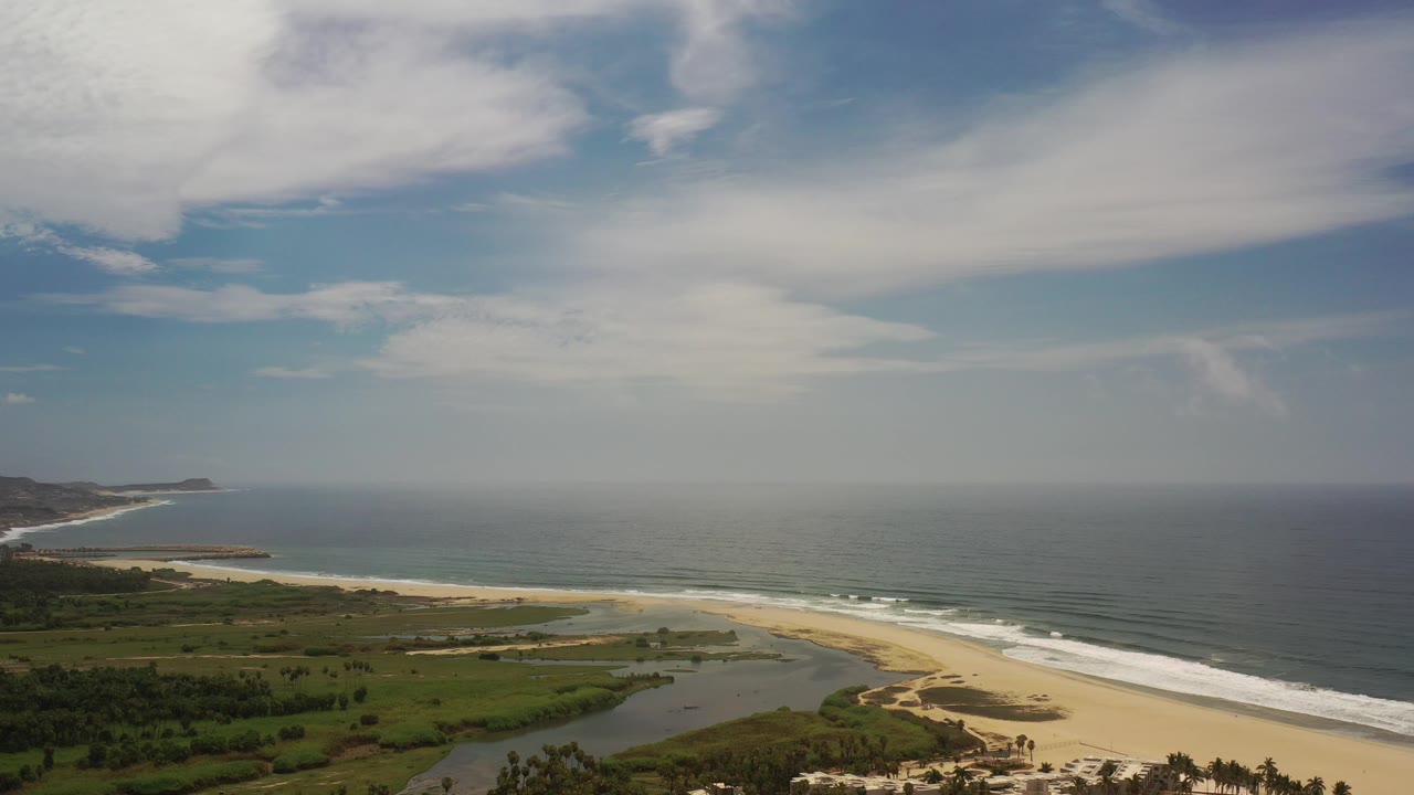 Drone-like coastline view with ocean, beach, lagoon, vegetation, and distant headland.