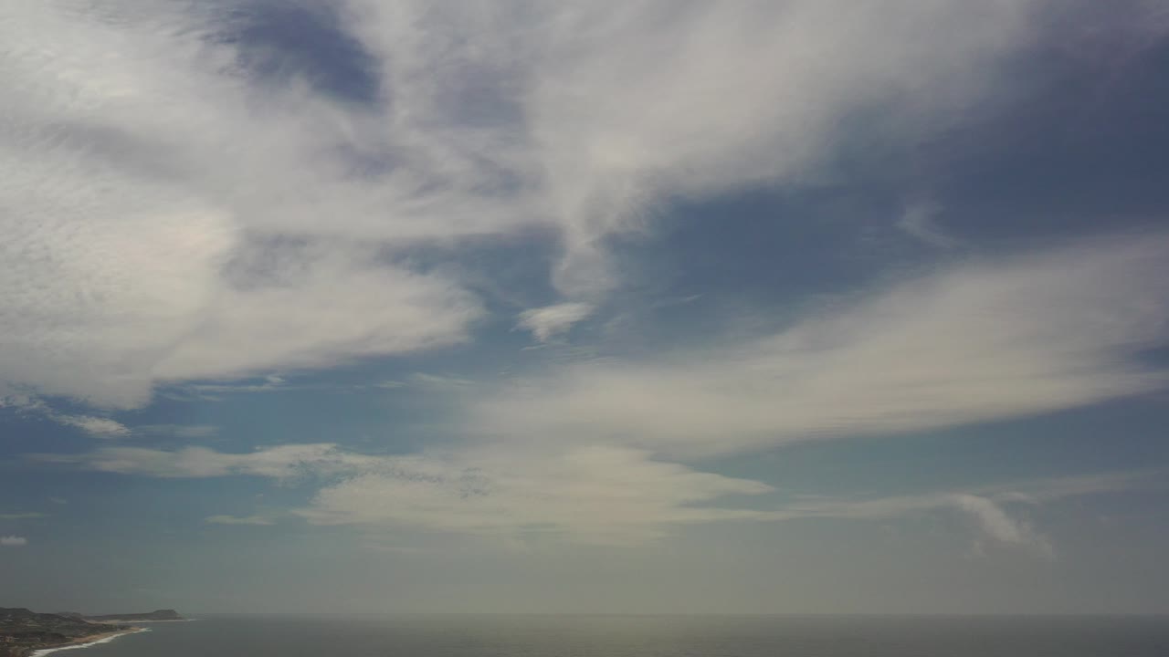 Wide view of blue sky and layered white clouds over a low ocean horizon in Los Cabos, Mexico, with a faint coastline at lower left and no visible people.