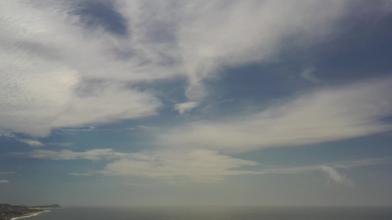 Blue sky and thick white cloud bands stretch over a hazy ocean horizon in Los Cabos, Mexico, with no visible people.