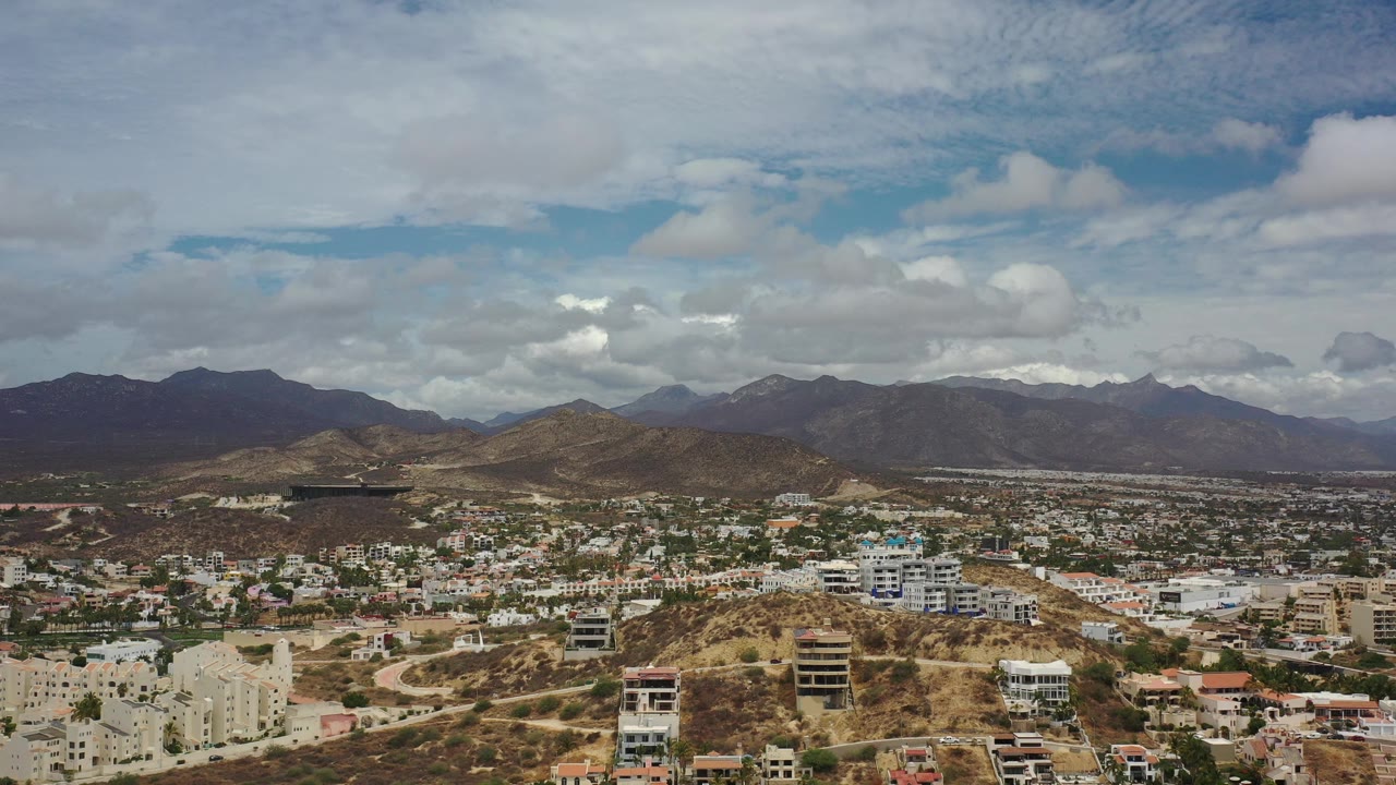 Opening aerial view over Los Cabos, Mexico, with tan hillside buildings, scattered white homes, distant mountain ridges, layered clouds, and no visible people.