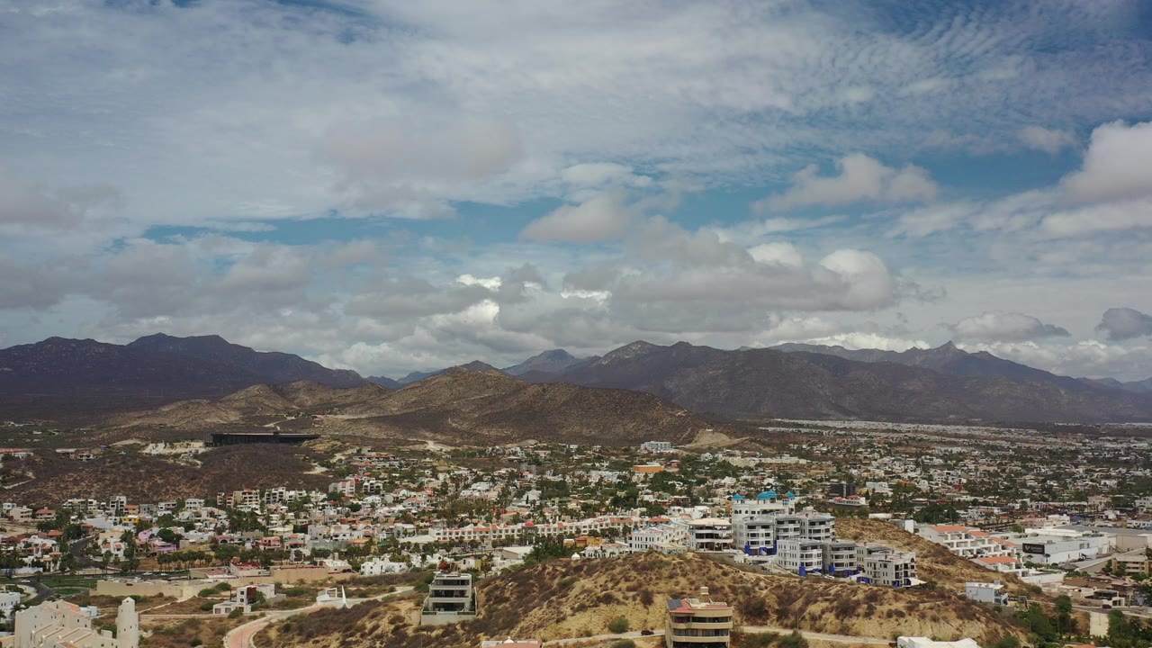 Opening aerial frame over Los Cabos showing low-rise buildings below dry hills, distant mountains, layered clouds, and no visible people.